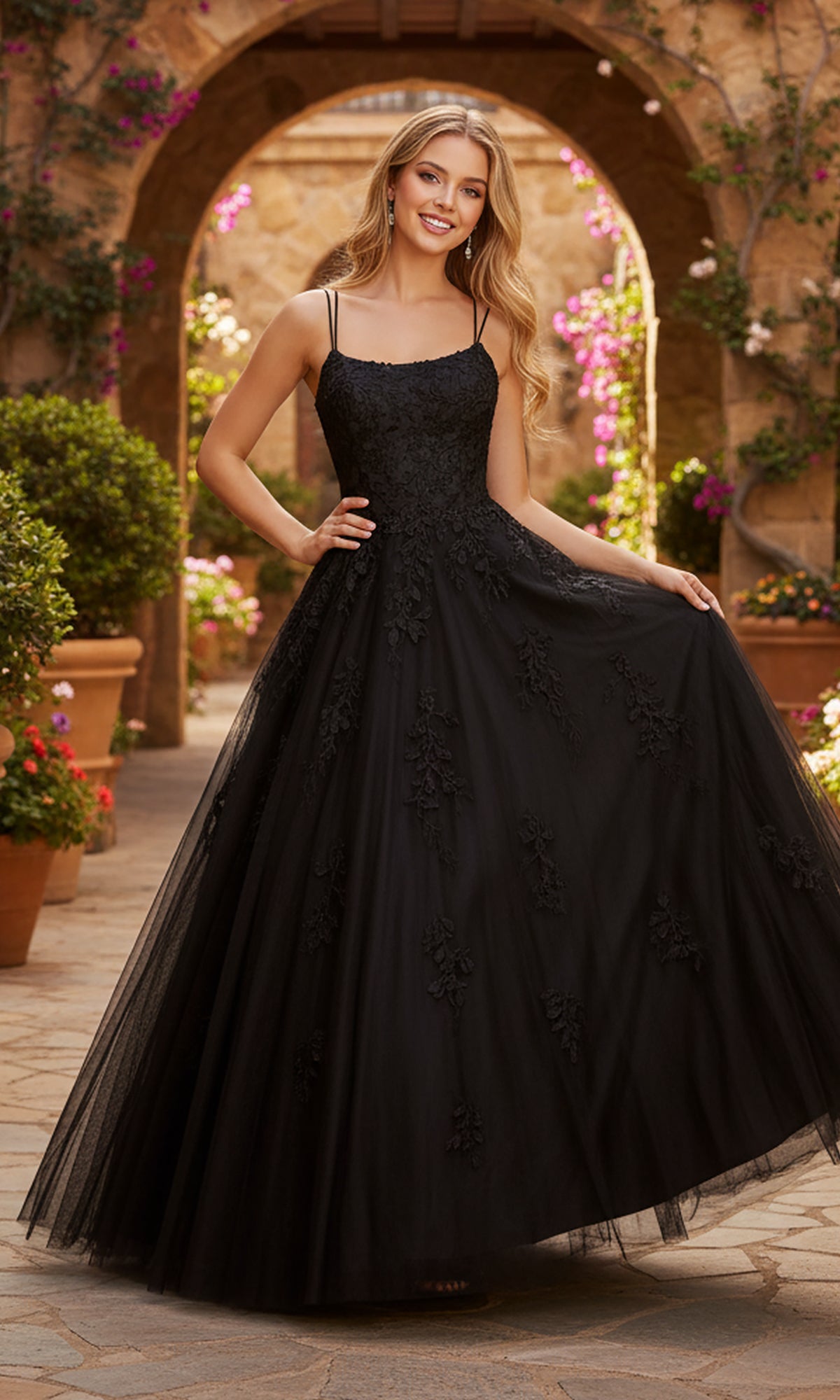 A woman smiles at the camera outdoors, wearing the PromGirl Exclusive 250228 long black lace-embroidered prom dress with thin straps and an adjustable lace-up back, holding the skirt out to each side.